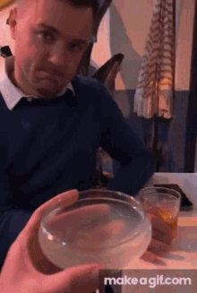 a man in a blue sweater is sitting at a table holding a glass of liquid in front of him