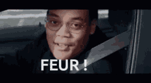 a man wearing glasses and a seat belt is sitting in a car and saying peur !