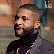 a man with a beard is wearing a suit and a turtleneck and a be it logo is behind him