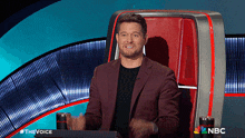 a man in a suit sitting in front of a nbc logo