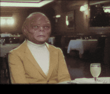 a man in a yellow jacket and white turtleneck sits at a table with a glass of wine
