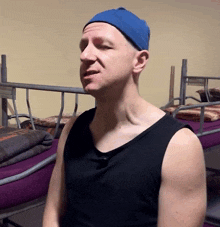 a man wearing a blue hat and a black tank top is standing in front of bunk beds