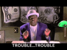 a man in a purple suit and hat is sitting at a desk with a green lamp and a sign that says trouble .