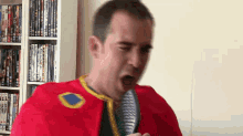 a man in a red cape is singing into a microphone in front of a bookshelf full of dvds