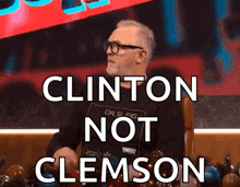 a man wearing glasses is sitting in front of a sign that says " clinton not clemson "