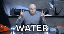 a bald man is sitting at a desk giving a peace sign and the word water is behind him .